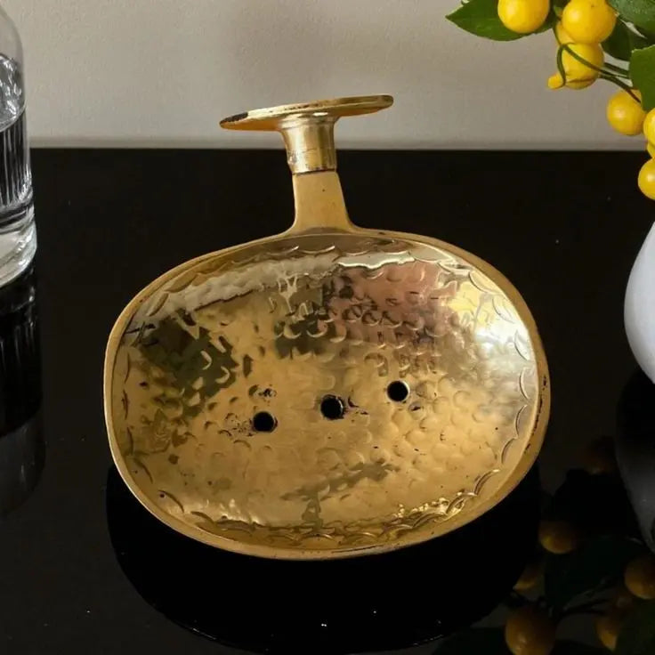 A golden, handcrafted unlacquered brass soap dish, either engraved or hammered, resting on a marble bathroom vanity.